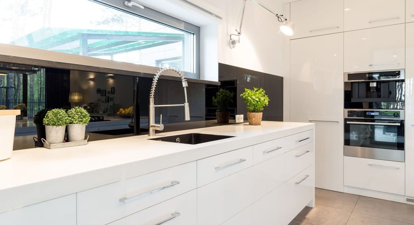 A modern kitchen with silver extendable silver tapware, white counter top, wall mounted oven, and large white storage spaces.
